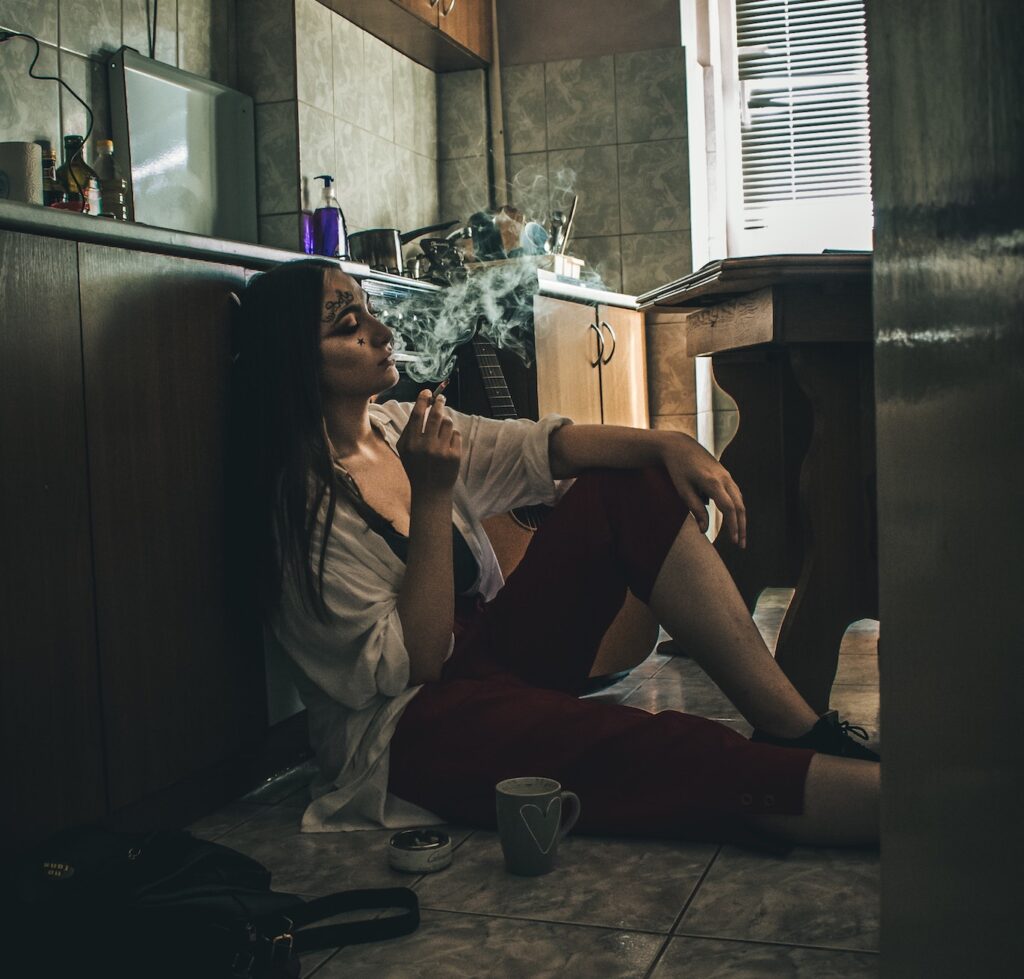Woman sits on floor of kitchen, smoking a cigarette.