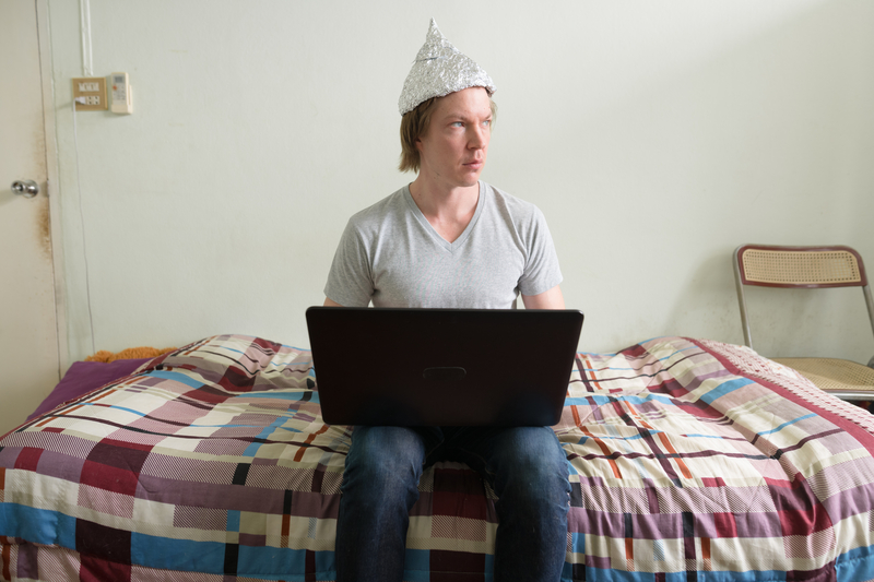 Man with tin foil hat sits on bed with laptop computer, looking to the side.