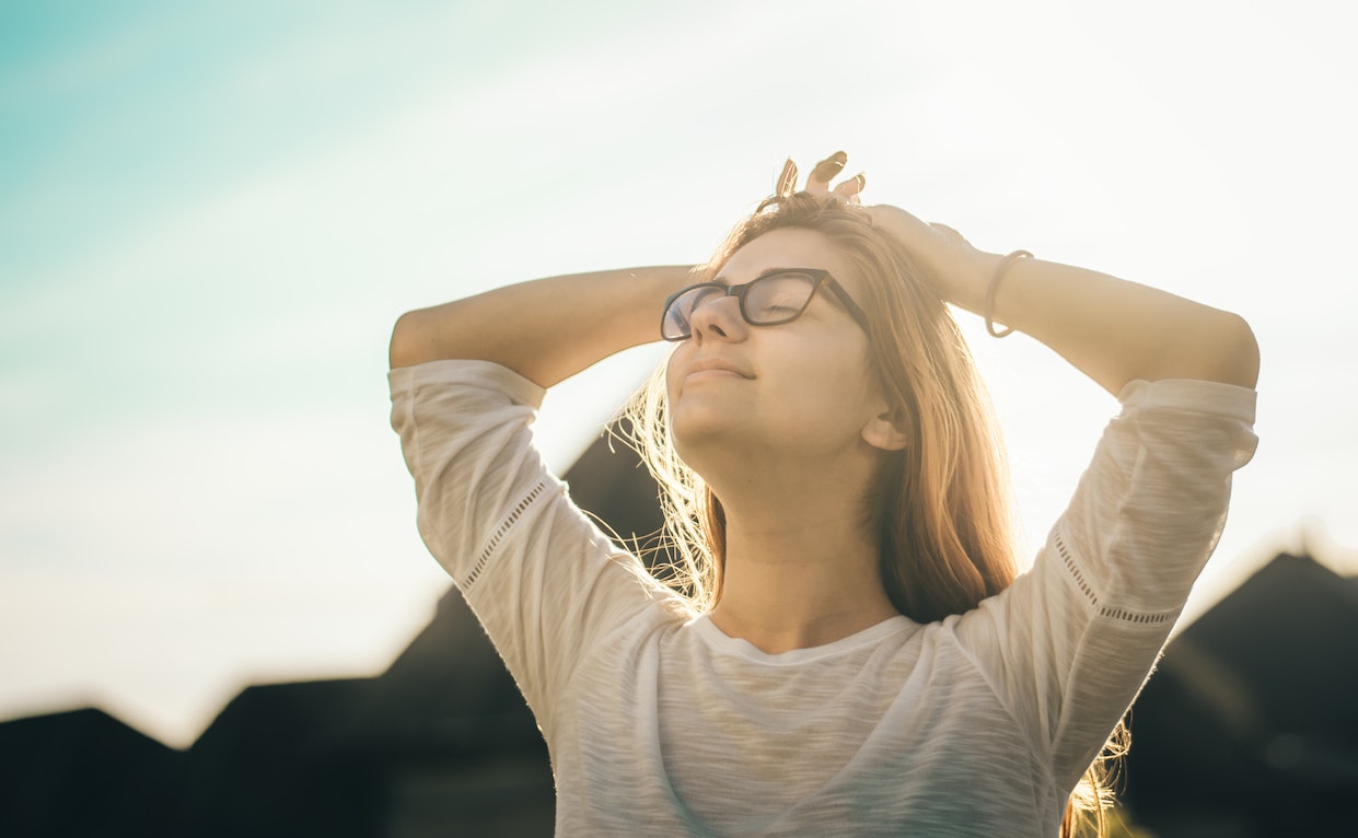 Woman standing outside, hands above head, looking up with eyes closed; happy
