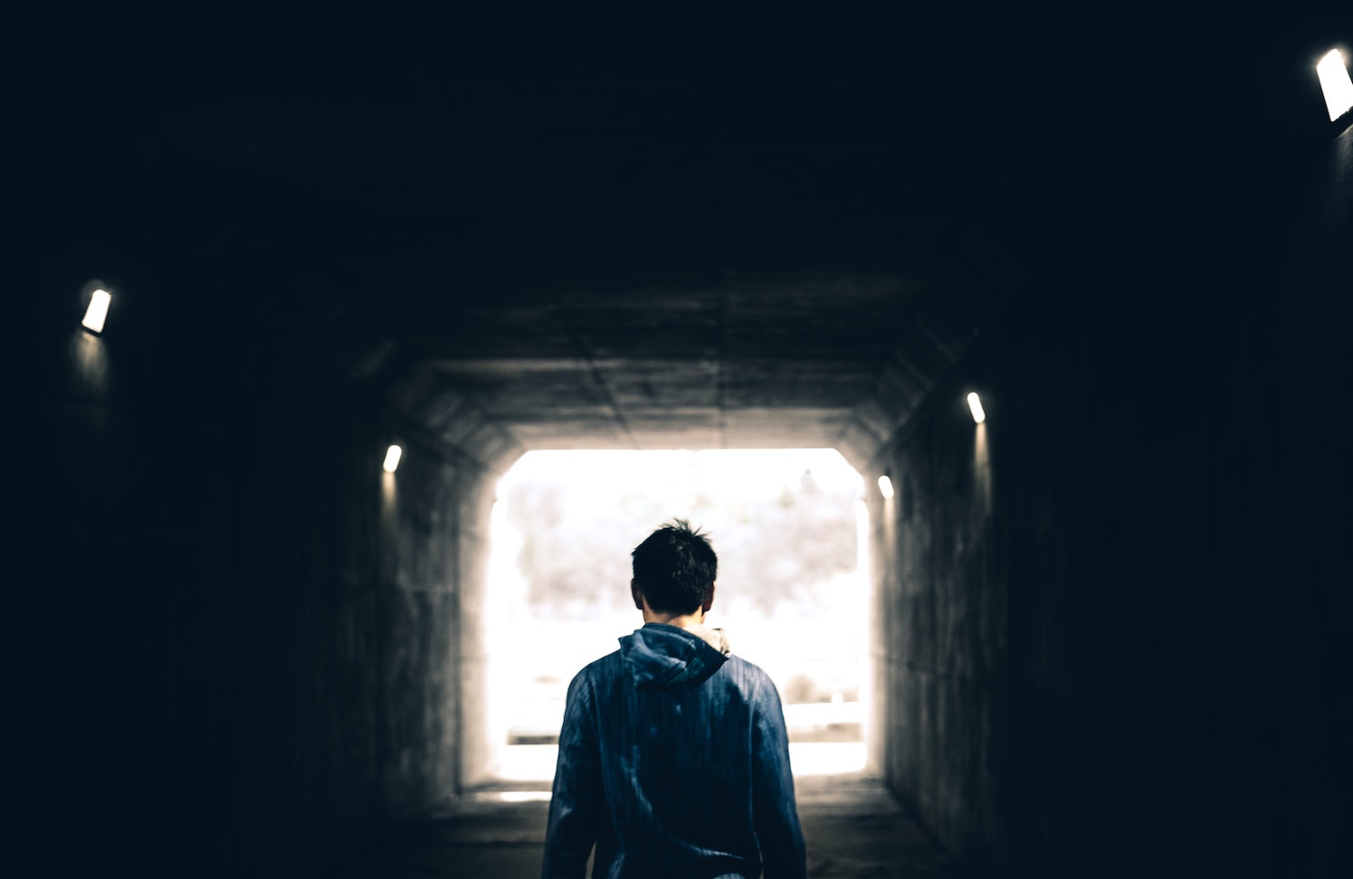 Black and white image of back of man standing, silhouetted by light at end of tunnel.