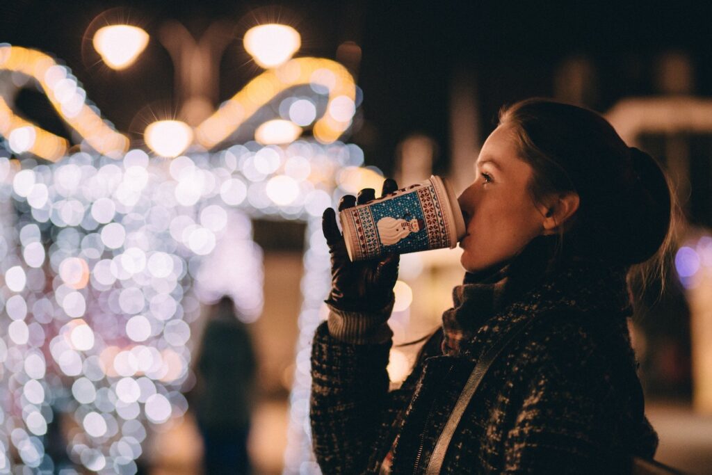 Woman drinking coffee from a holiday coffee cup outside in front of Christmas lights