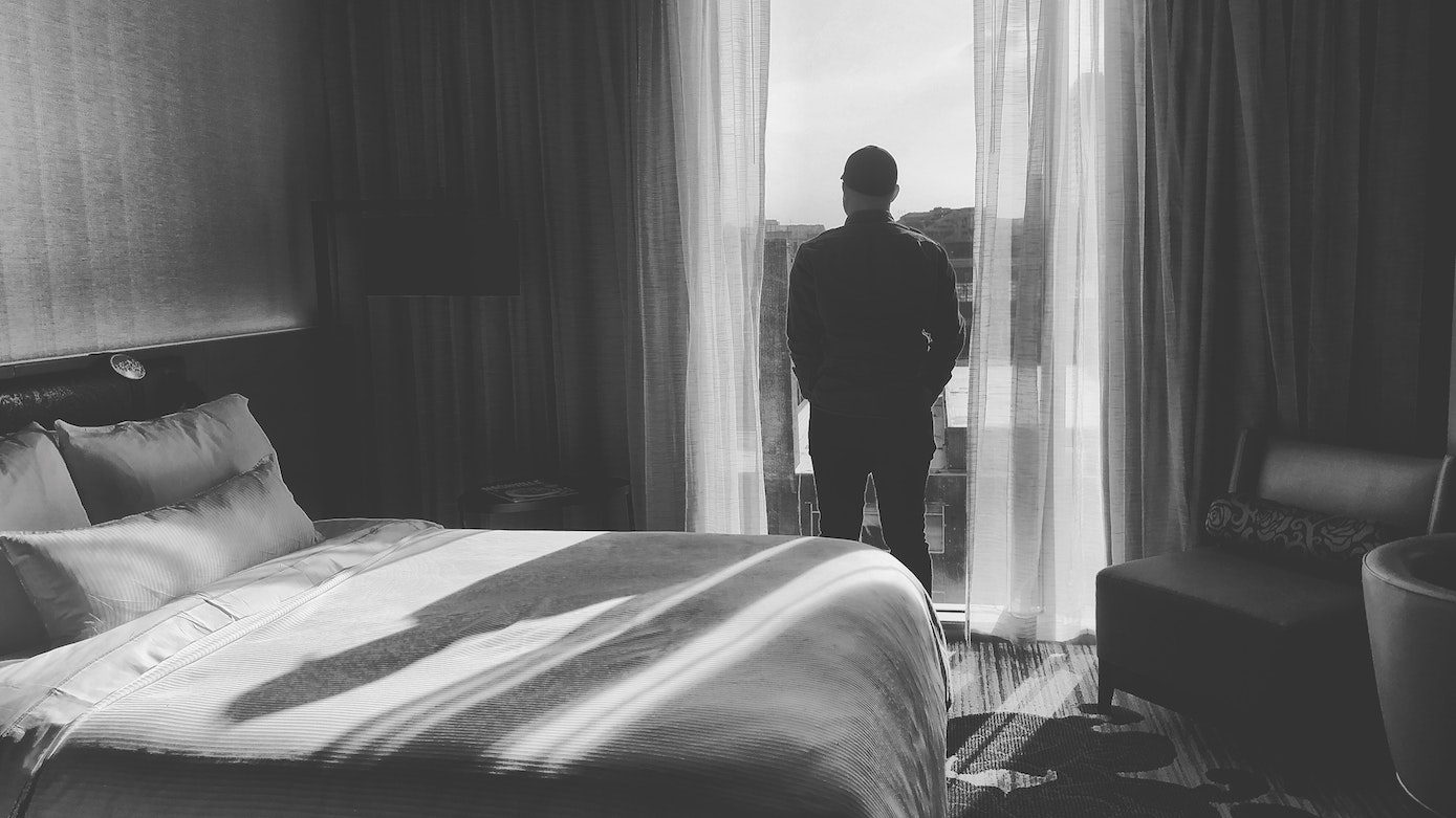 Black and white image of bedroom, view of back of man standing looking out window