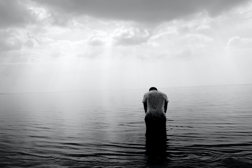 Black and white image of man, from back, kneeling in water under cloudy sky