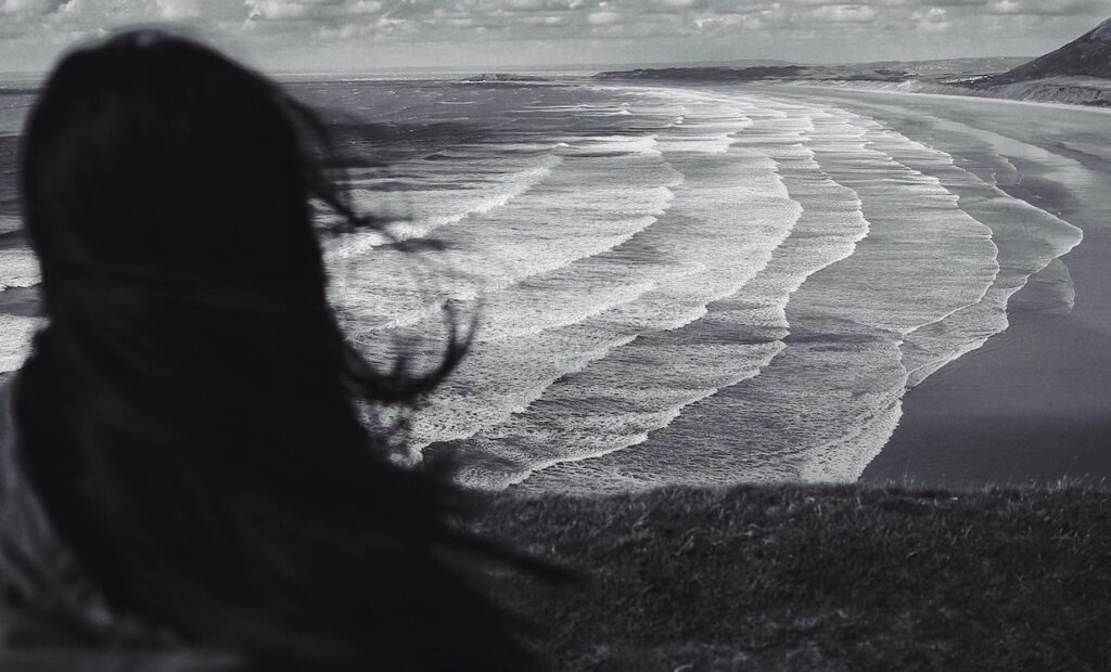Return to Sender: What an Unsent Postcard Taught Me About Addiction Black and white image of woman from back looking over waves breaking on the sand