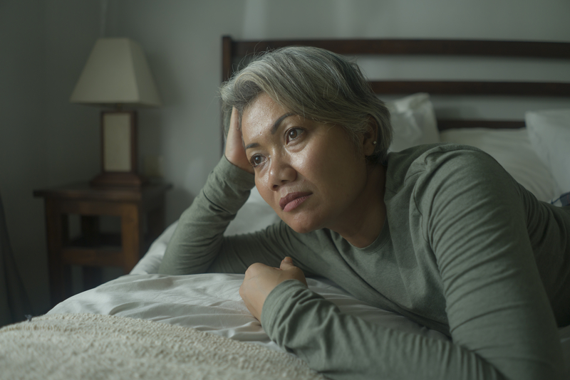 Woman with gray hair on bed, head resting on hand, sad.