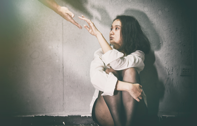 A sad woman sits against a wall, reaches hand up to accept outstretched helping hand