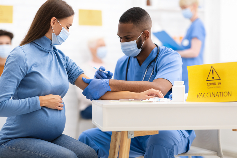 Masked pregnant woman gets the covid vaccine from masked health worker in a medical setting.