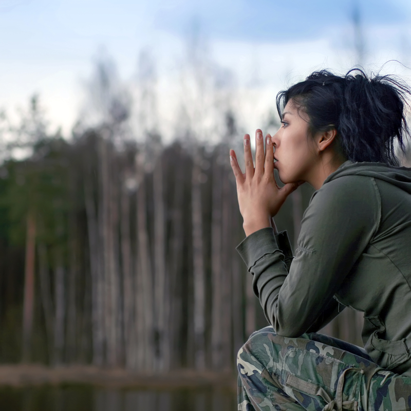 Woman with dark hair and camouflage pants sitting outside, in contemplation