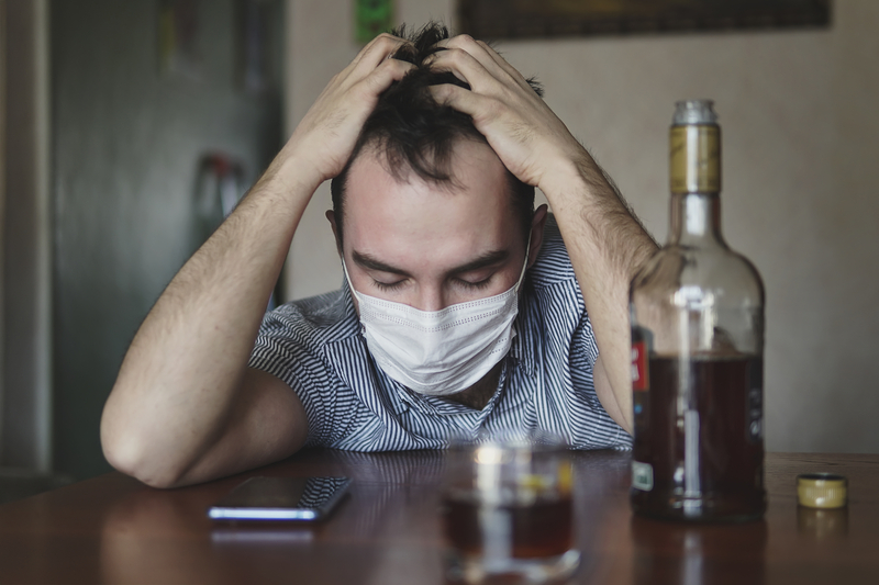 Man in mask sits at bar with alcohol bottle and glass, head in hands.