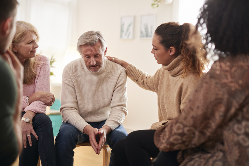 Man in group therapy looks sad. Woman next to him rests her hand on his shoulder.
