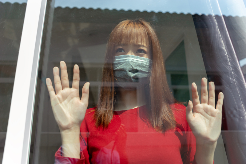 Woman with mask stands looking out window, hands against the glass