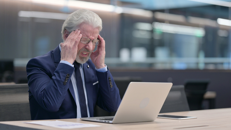 Man holding head in hands, headache, sitting at table with computer