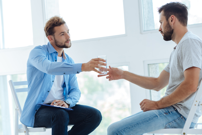 Clinician offers water to patient, both sitting in bright room with windows.